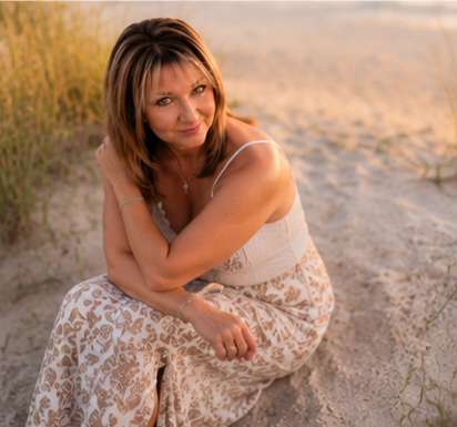 Woman in floral maxi skirt sitting on sandy beach at golden hour with soft dunes and grass in background.