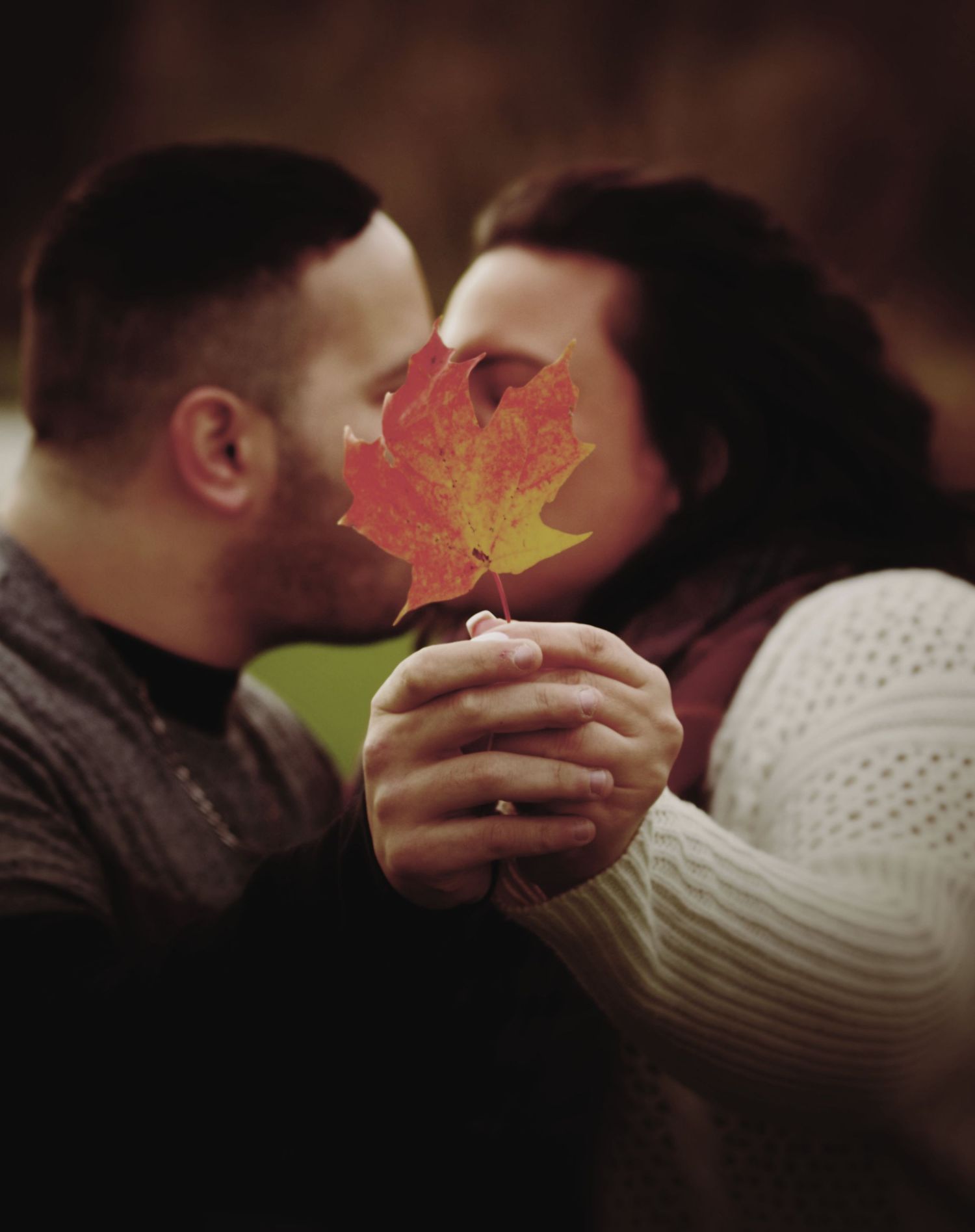 Couple kissing behind a vibrant red autumn maple leaf held in their hands during a fall outdoor photo session.