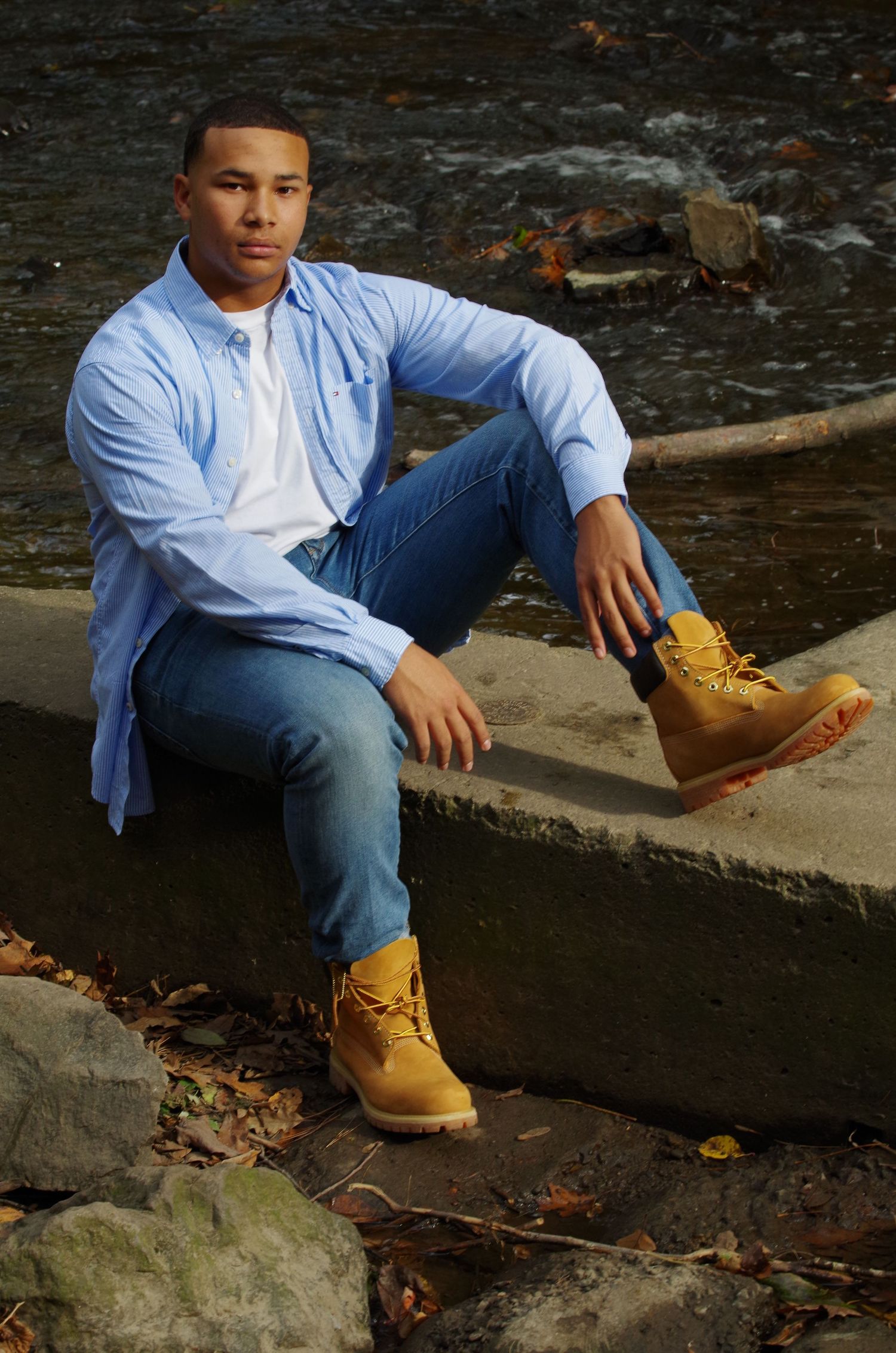 Person in light blue shirt and jeans sitting casually on stone ledge near water.