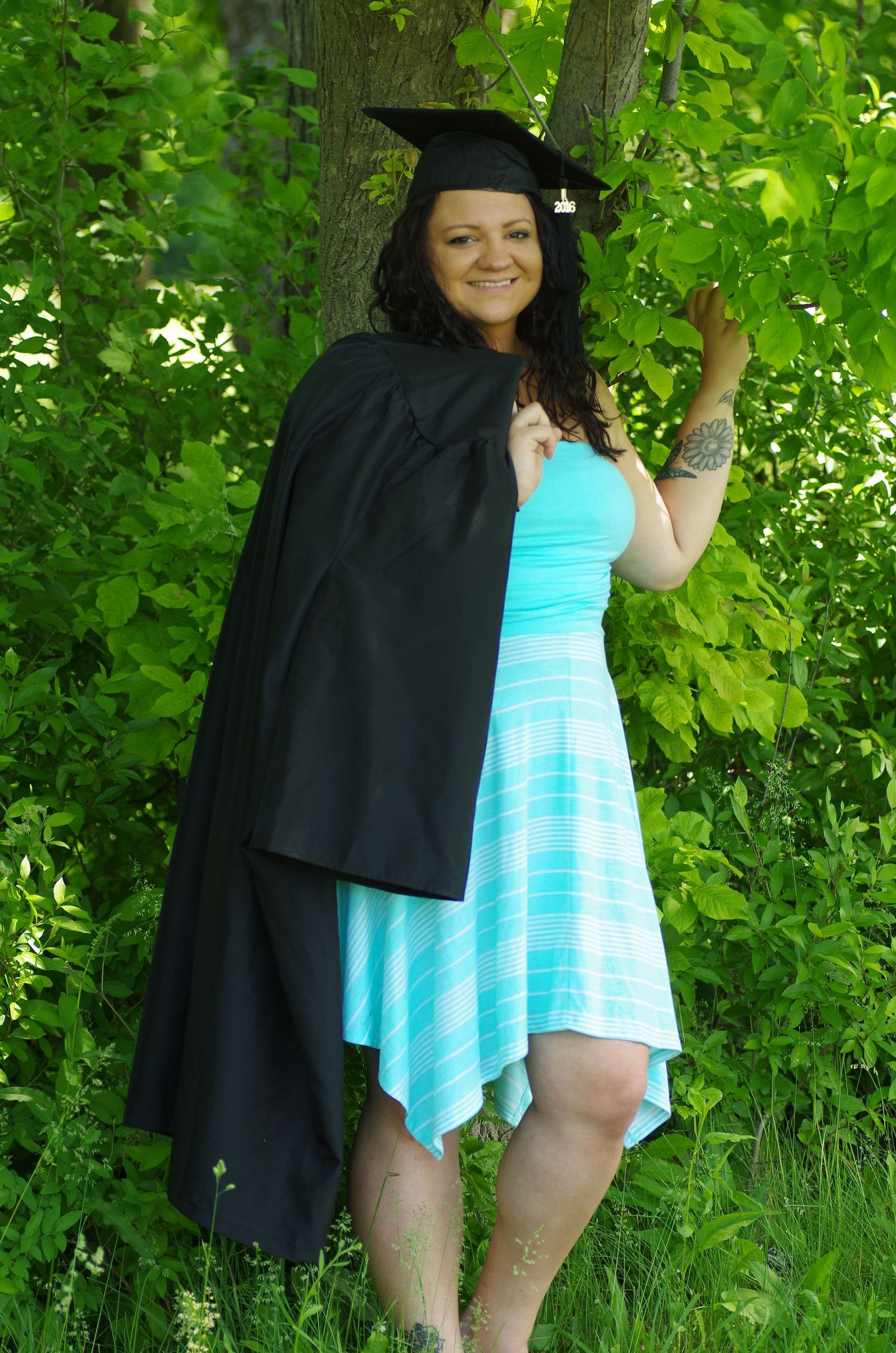 Graduate in black cap and gown with turquoise dress poses playfully against green foliage.