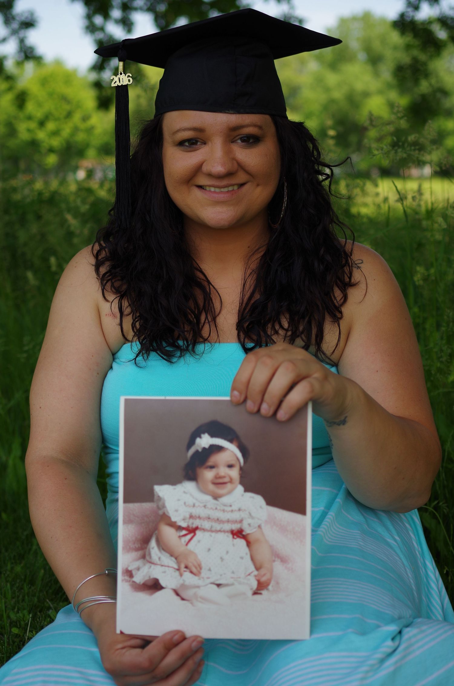 Graduation photo showing graduate holding baby photos while seated outdoors.