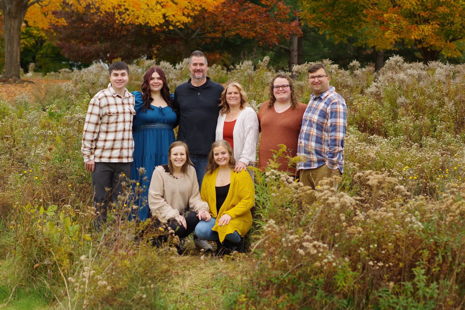Group portrait of eight people posing outdoors among tall grass and fall foliage.