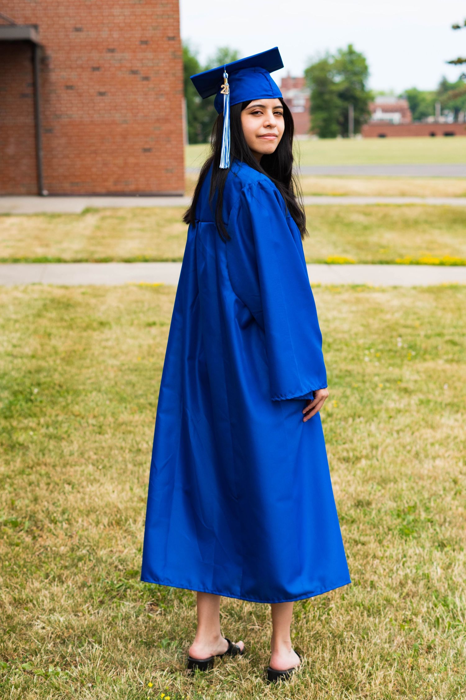 Graduate in blue cap and gown stands on grass near brick building.