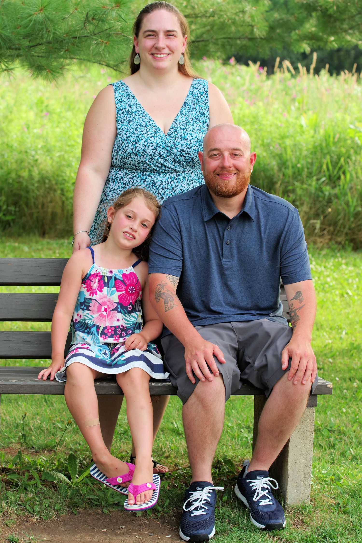 Family sits together on wooden park bench with green summer foliage in background.