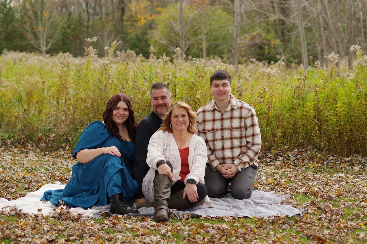 Small group sitting on blanket in field with tall grass and autumn foliage backdrop.