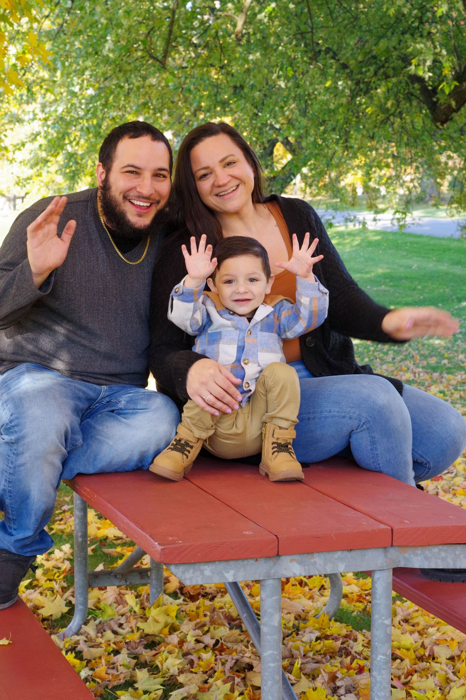 Family sits together on red picnic table surrounded by fall foliage in outdoor park setting.