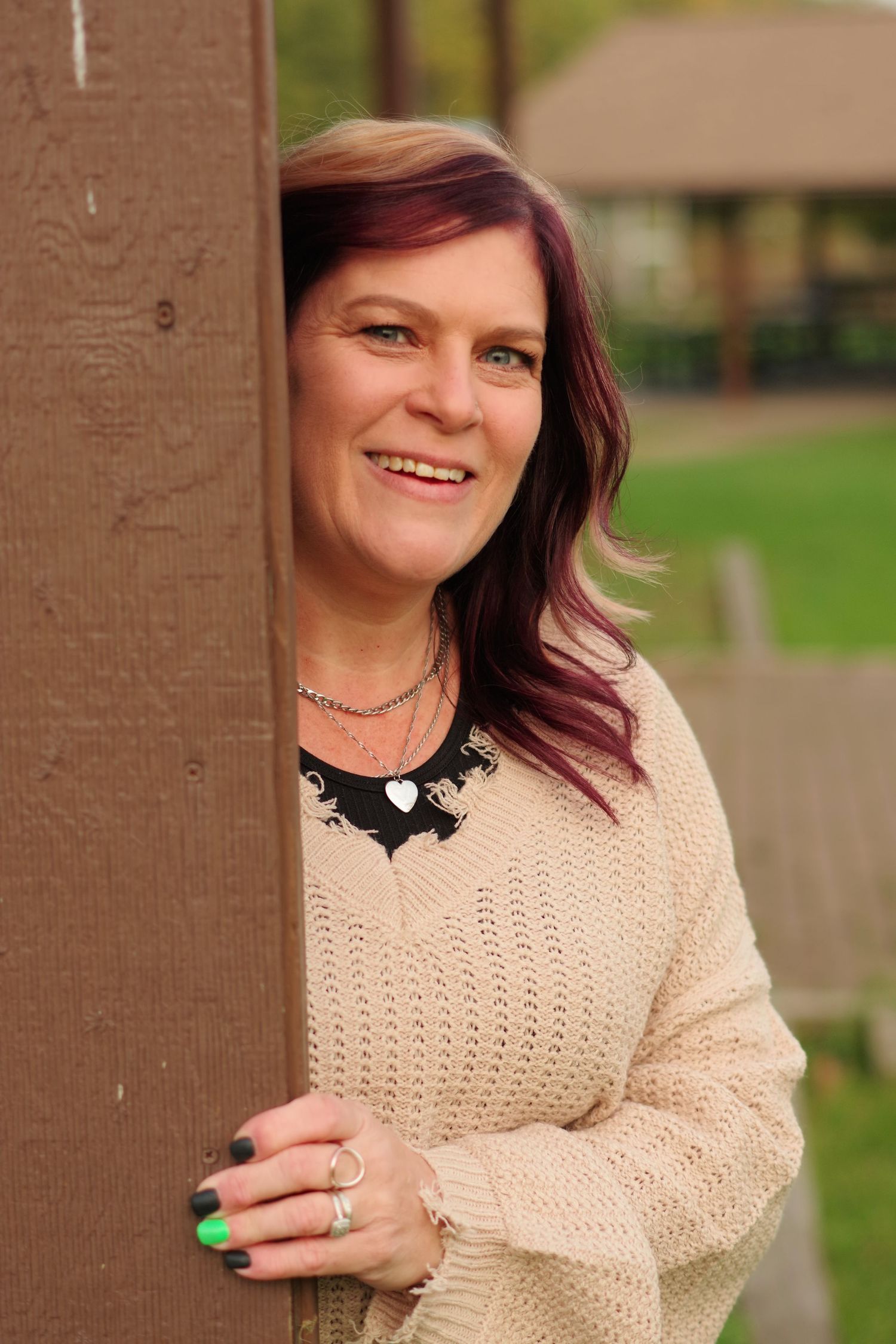 Person in cream sweater and statement necklace poses by wooden post with bright nail polish.