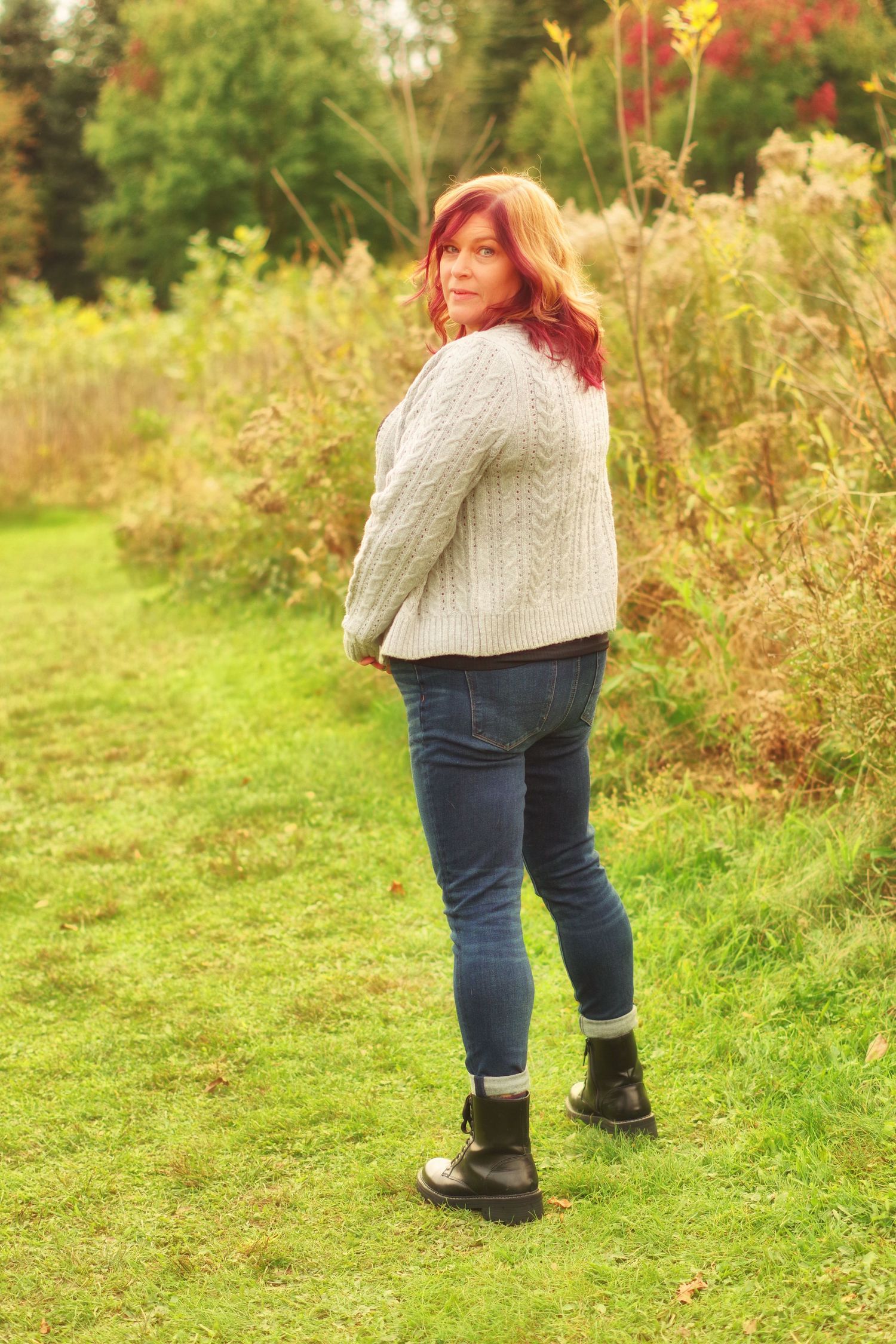 Side view portrait in natural setting wearing cream cable knit sweater and dark boots.