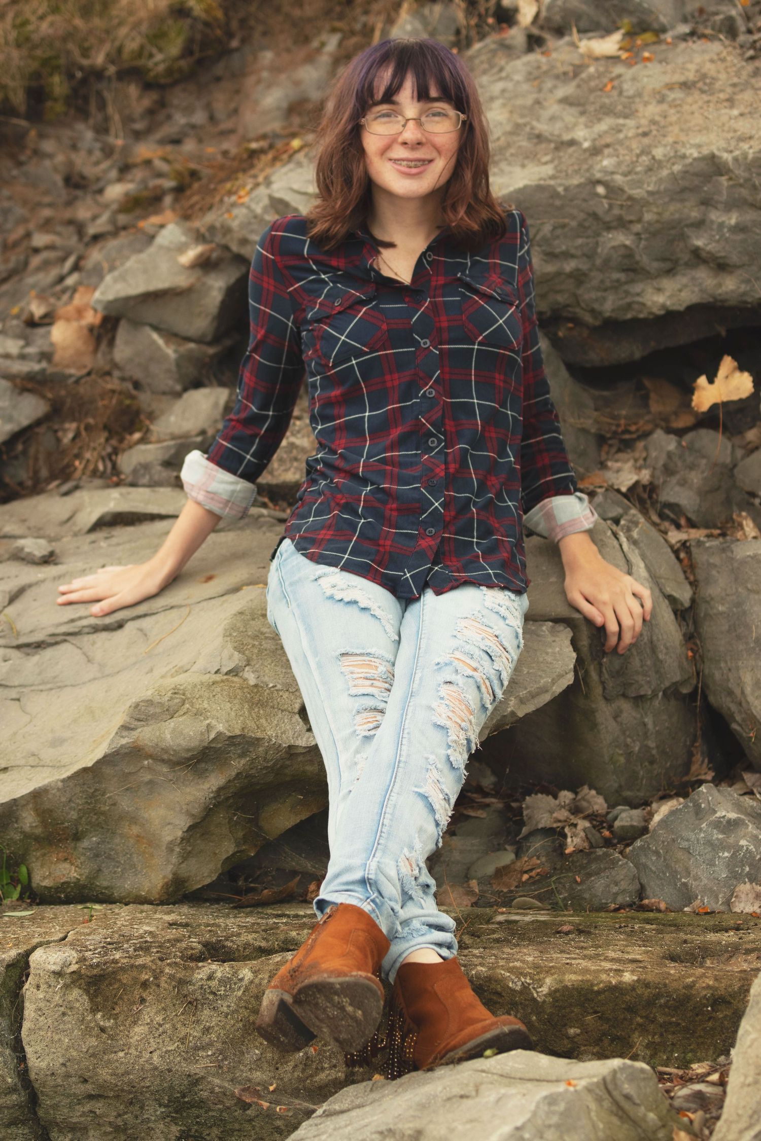 Person in plaid shirt and ripped jeans sitting on rocky terrain wearing brown boots.
