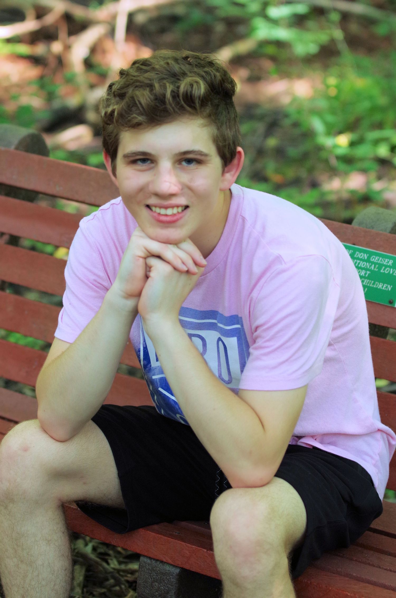 Someone in a pink t-shirt sits relaxed on a wooden park bench with hands folded.