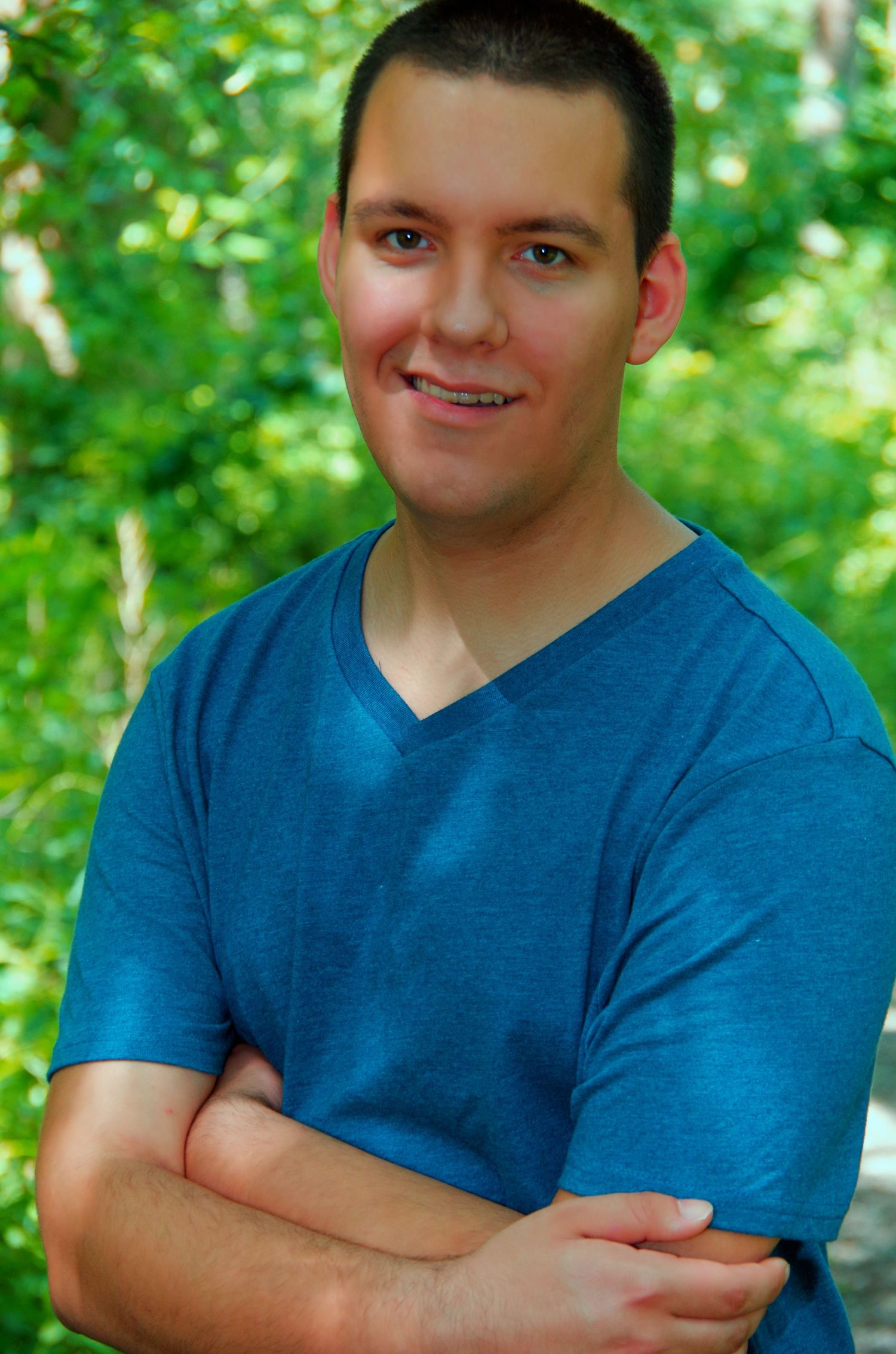A person in a blue v-neck t-shirt poses with crossed arms against green foliage.