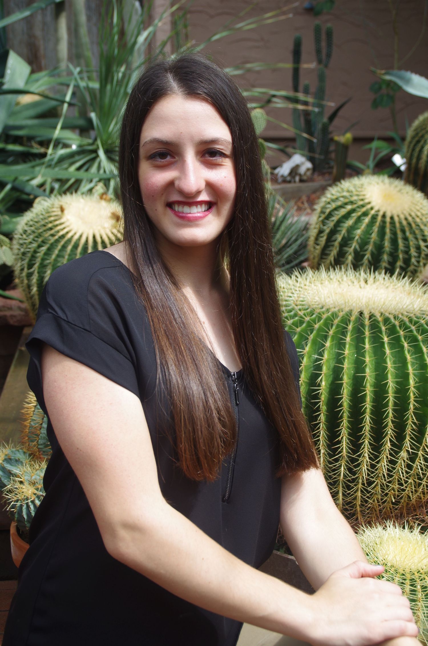 Person in black poses casually next to large barrel cacti in a greenhouse setting.