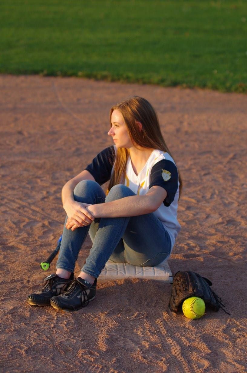 Softball player sits on dirt field at sunset with yellow ball and glove nearby.