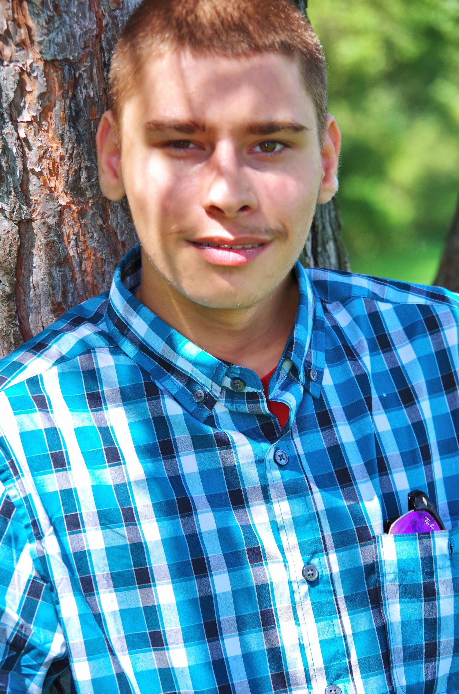 Someone in a blue and white checkered shirt smiling outdoors near a tree.