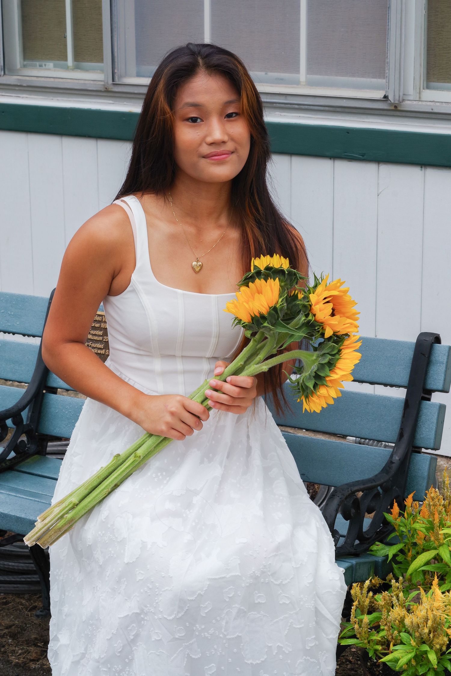 Person in white summer dress holding fresh sunflowers while standing on blue bench.