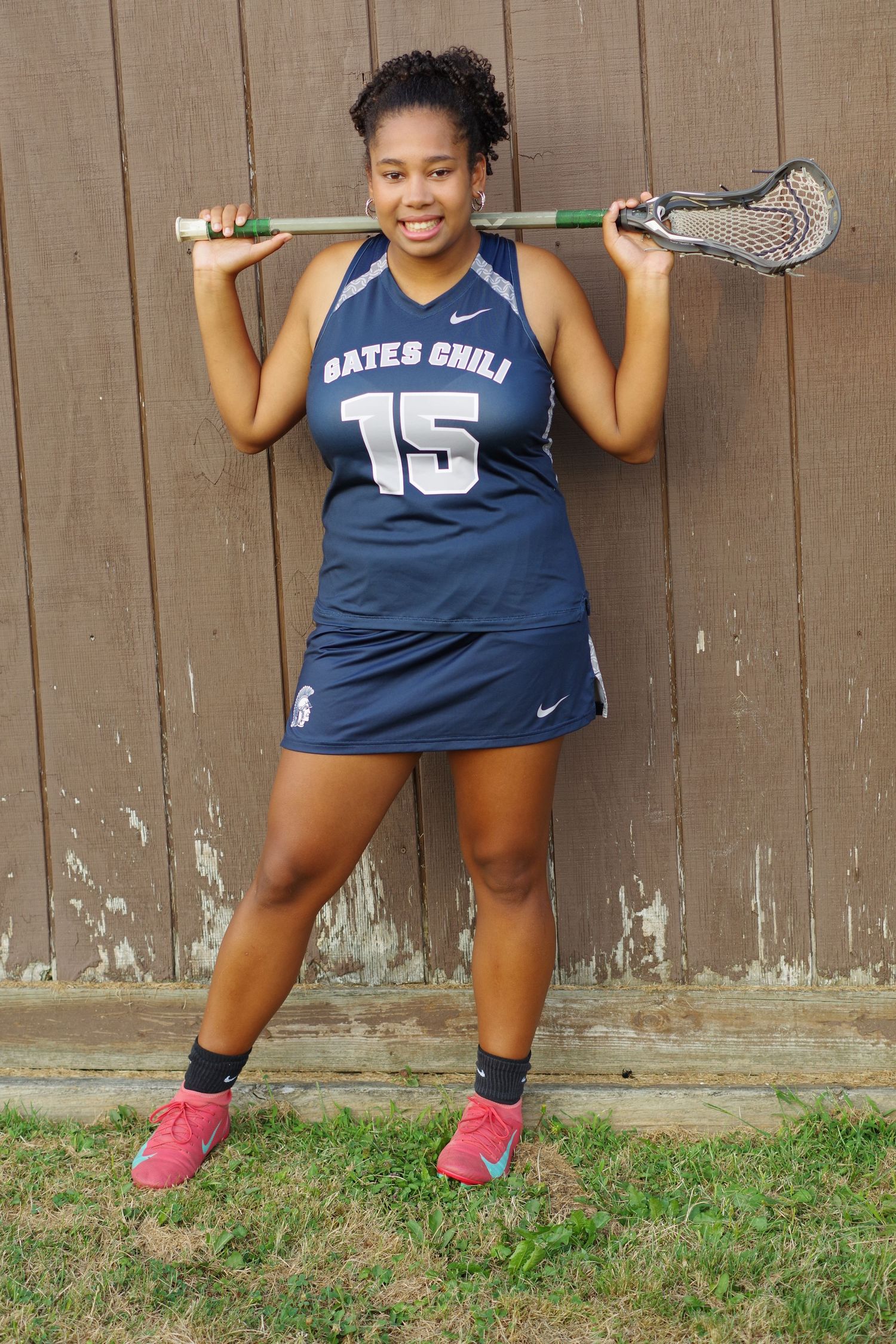 Athlete poses with lacrosse stick wearing navy Gates Chili #15 uniform against brown wall.