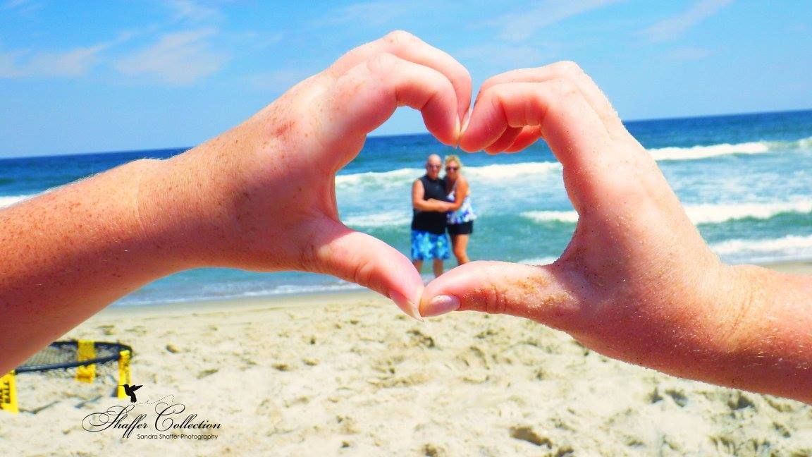 Hands forming heart shape framing distant beachgoers against ocean backdrop.