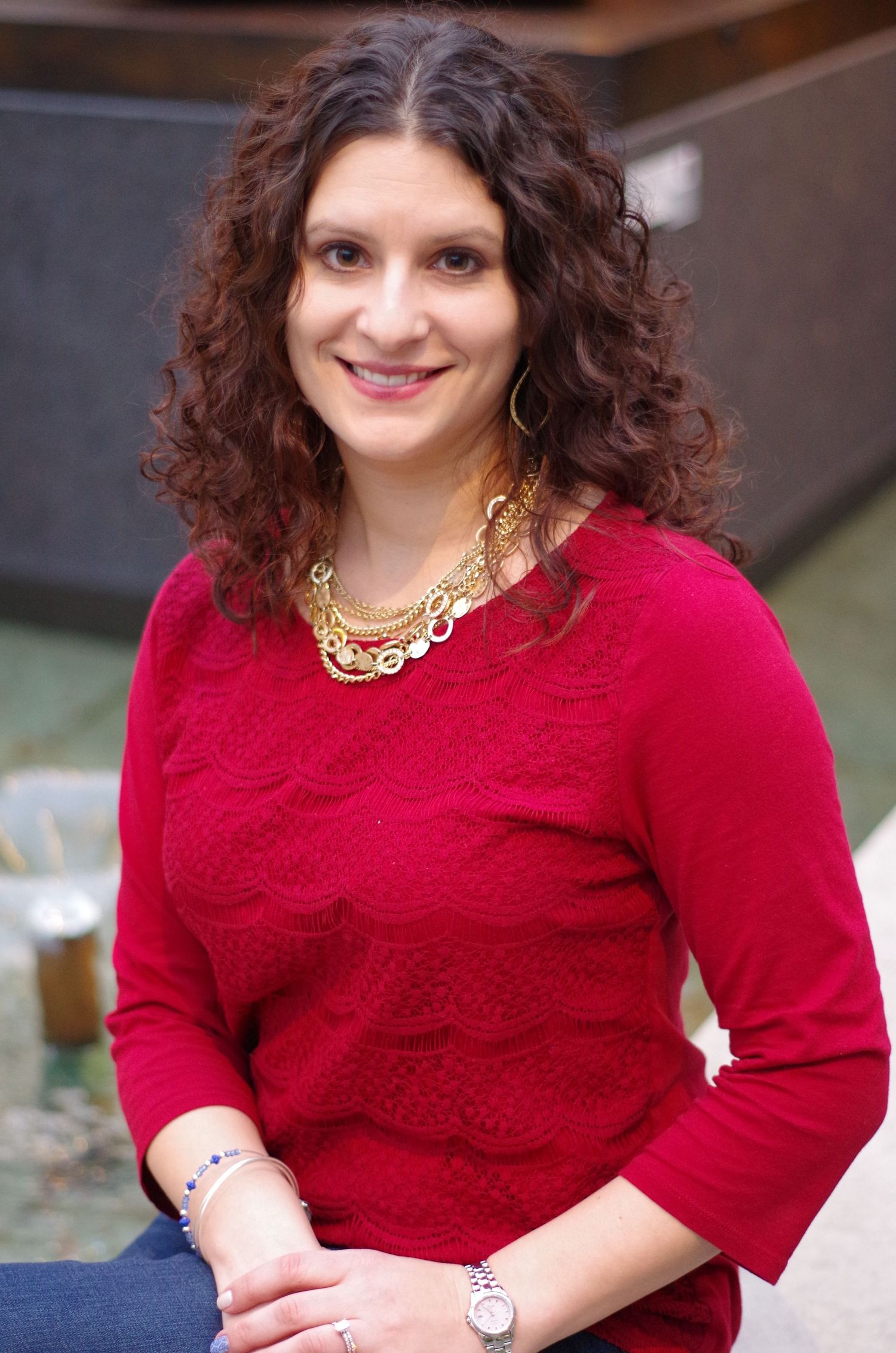 A person wearing a red sweater and statement necklace smiles at the camera.