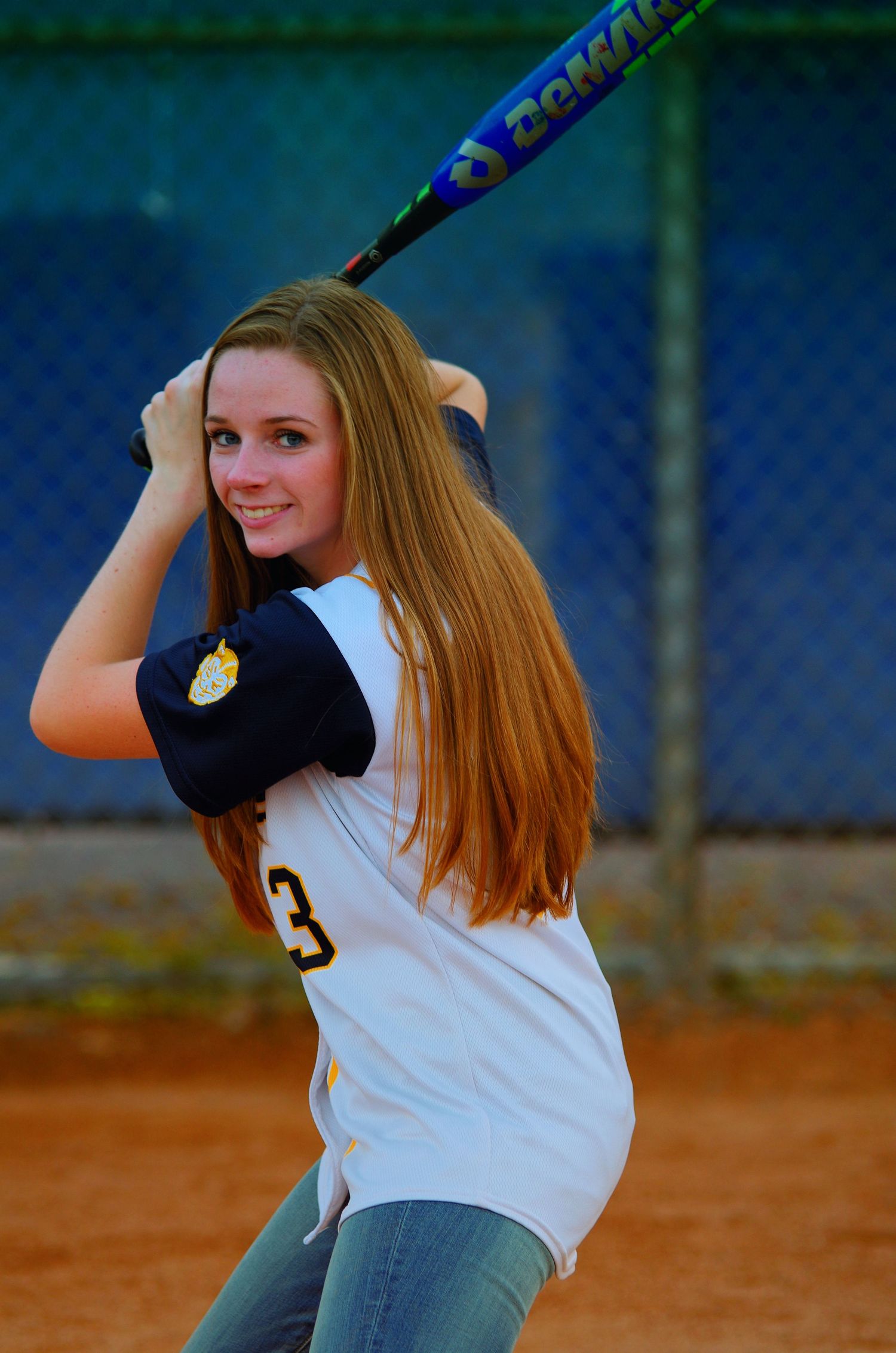 Softball player in uniform holds bat over shoulder on dirt field with blue fence background.