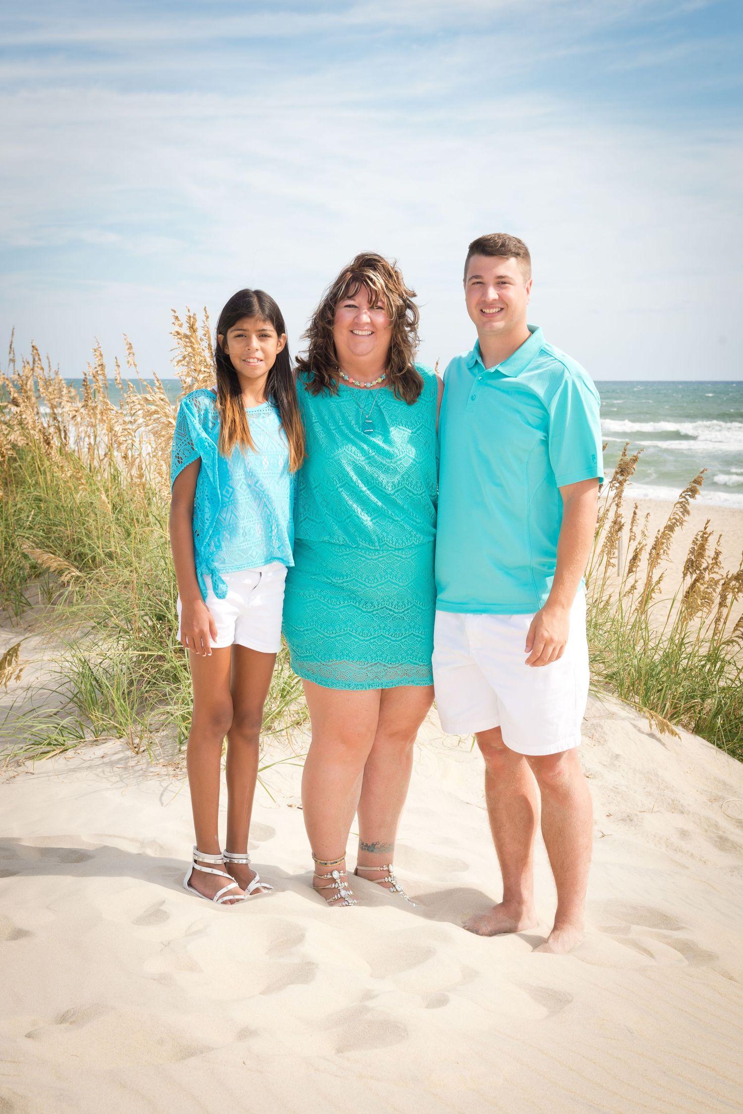 A family group in matching turquoise outfits poses together on a sandy beach with sea oats and ocean in background.