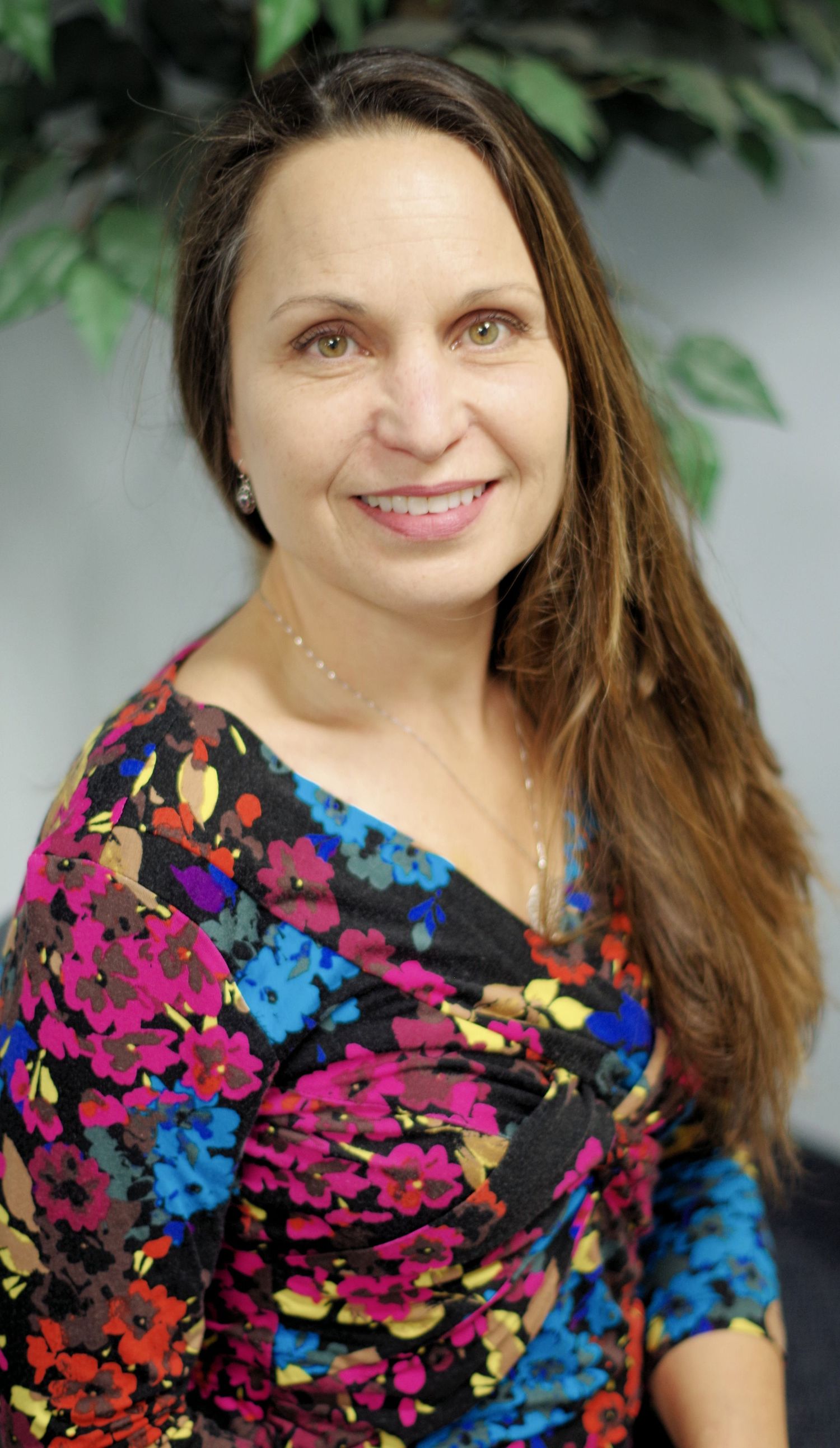 Portrait of a person wearing a colorful floral dress with long hair against a green plant backdrop.