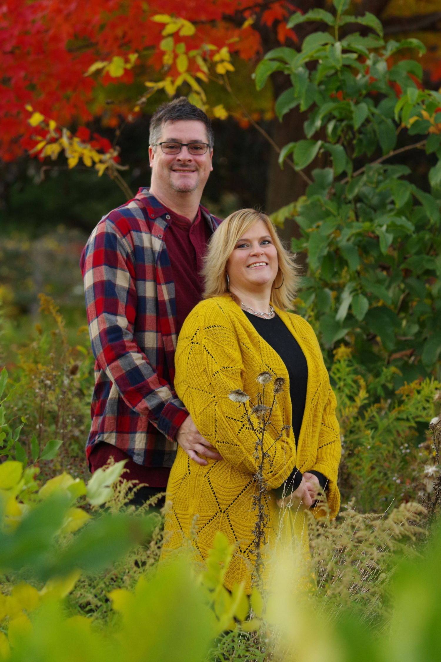 Couple poses outdoors in autumn with red foliage, wearing plaid shirt and yellow sweater.