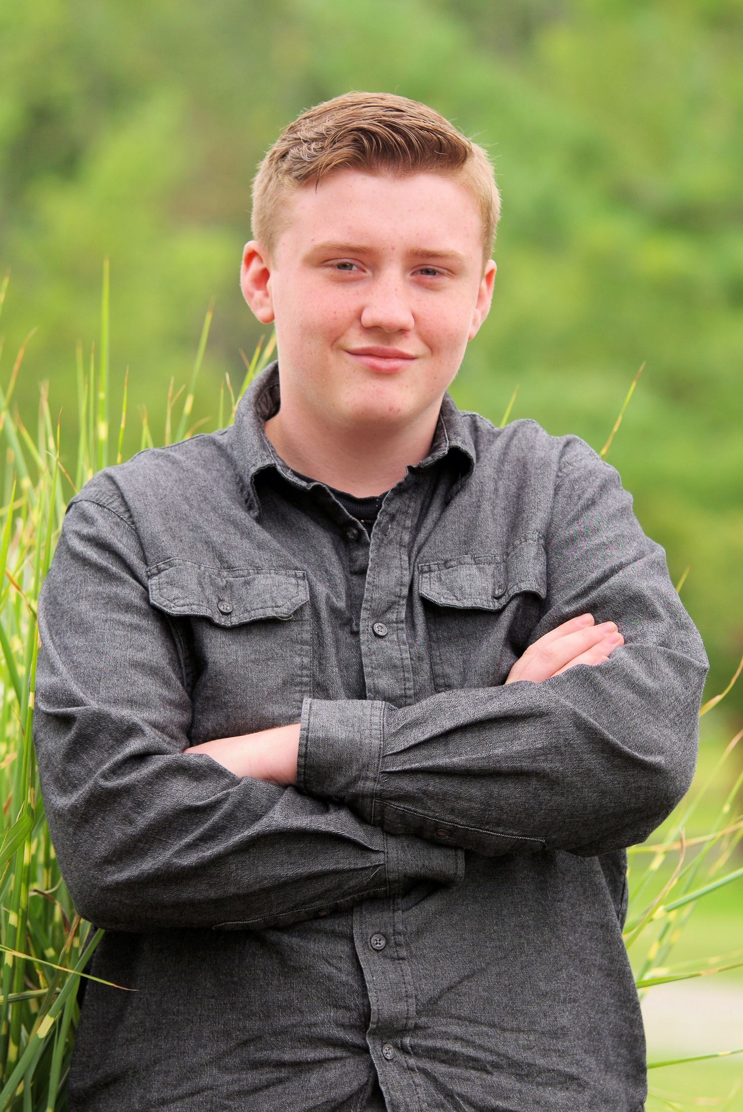 Person in gray work shirt poses with arms crossed against green grass background.