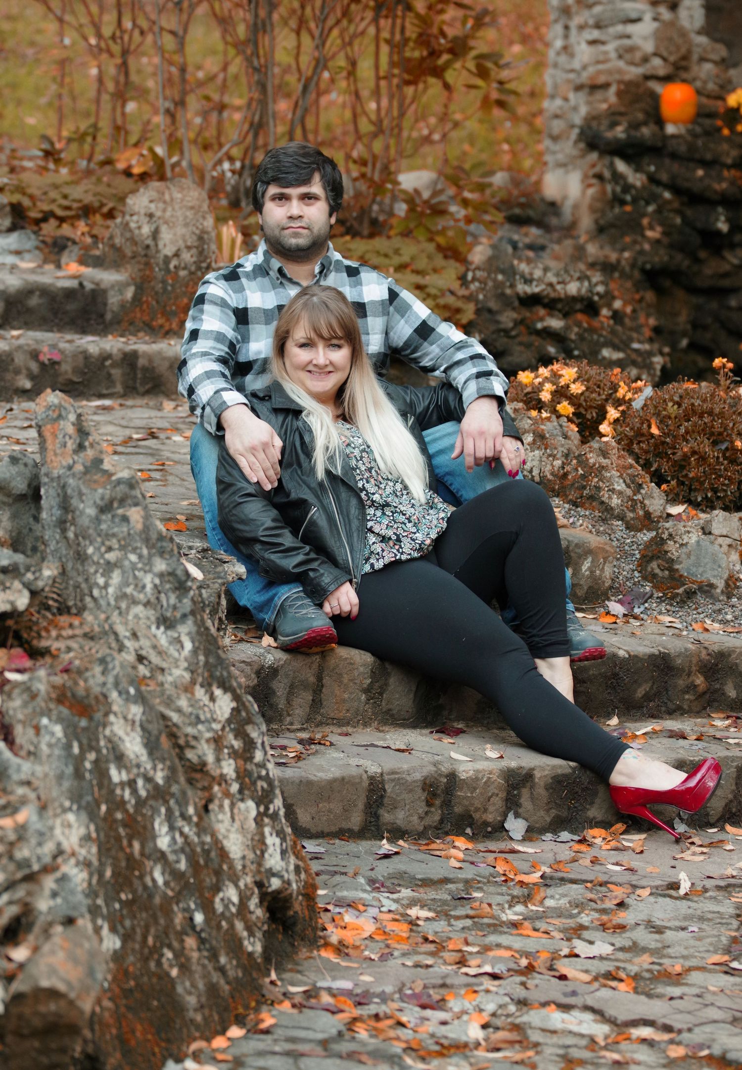 Couple sitting on stone steps surrounded by autumn foliage, woman in black outfit with red heels, man in plaid shirt.