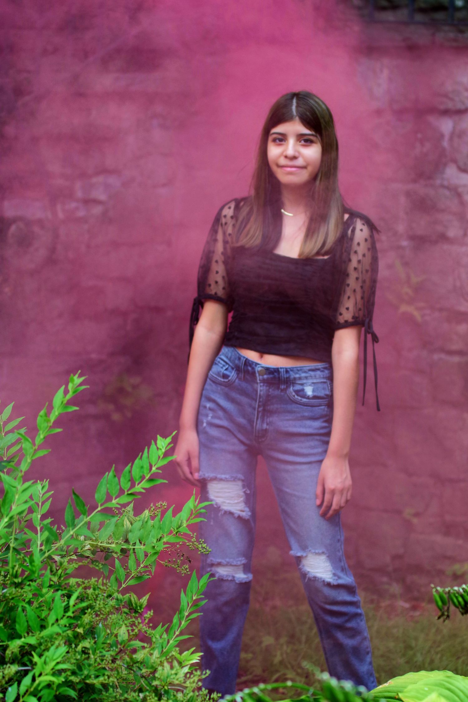 Young woman in black crop top and ripped jeans posing against a pink smoke-filled wall outdoors.