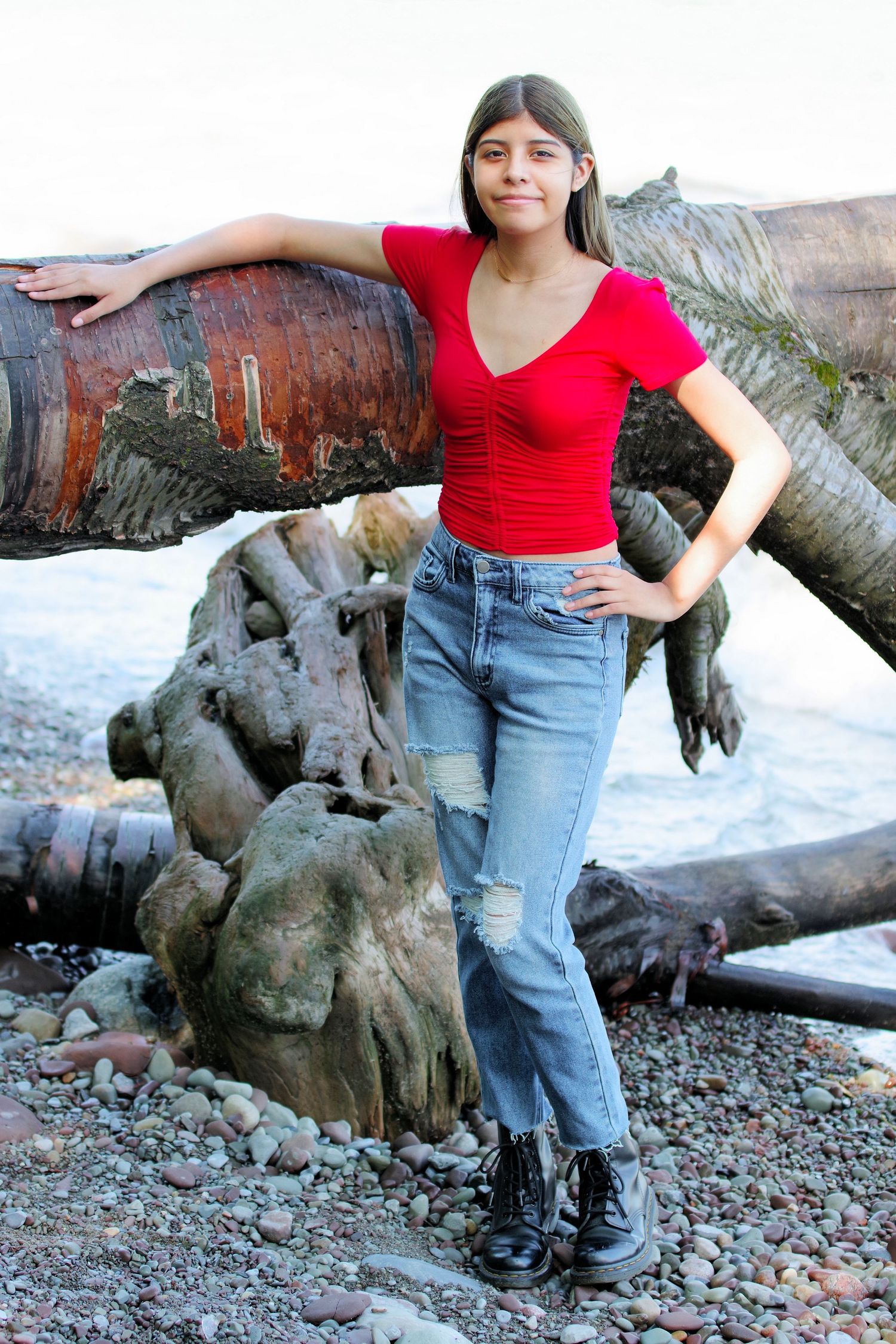 Portrait against driftwood logs on a rocky beach wearing red top and jeans with combat boots.