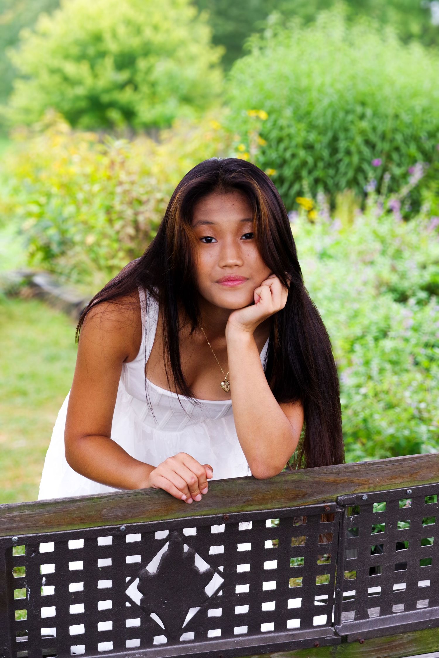 Person with long dark hair resting chin on hand while leaning on decorative wooden railing in garden setting.
