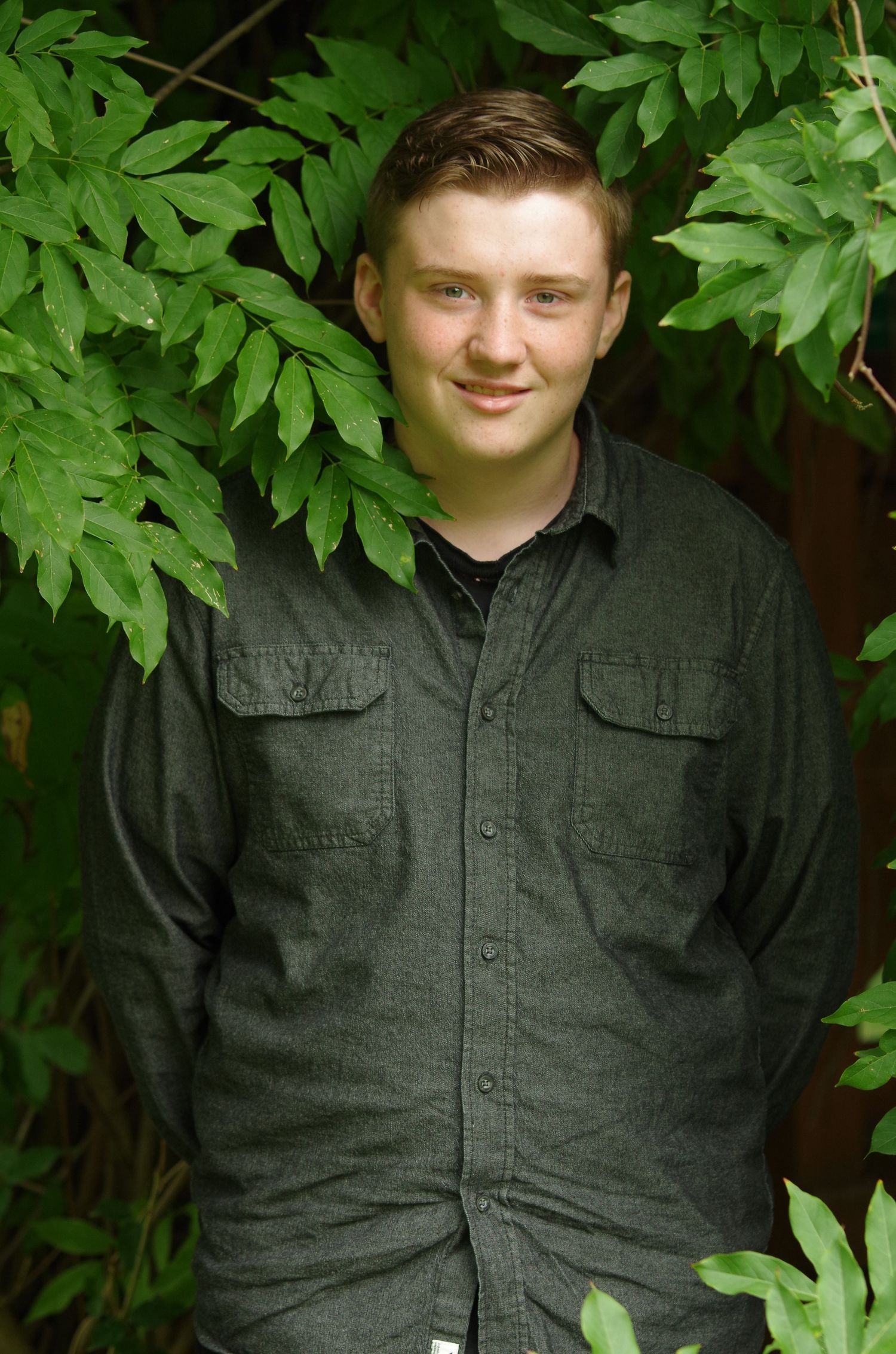 Portrait shot against lush green leaves wearing a dark button-up shirt.
