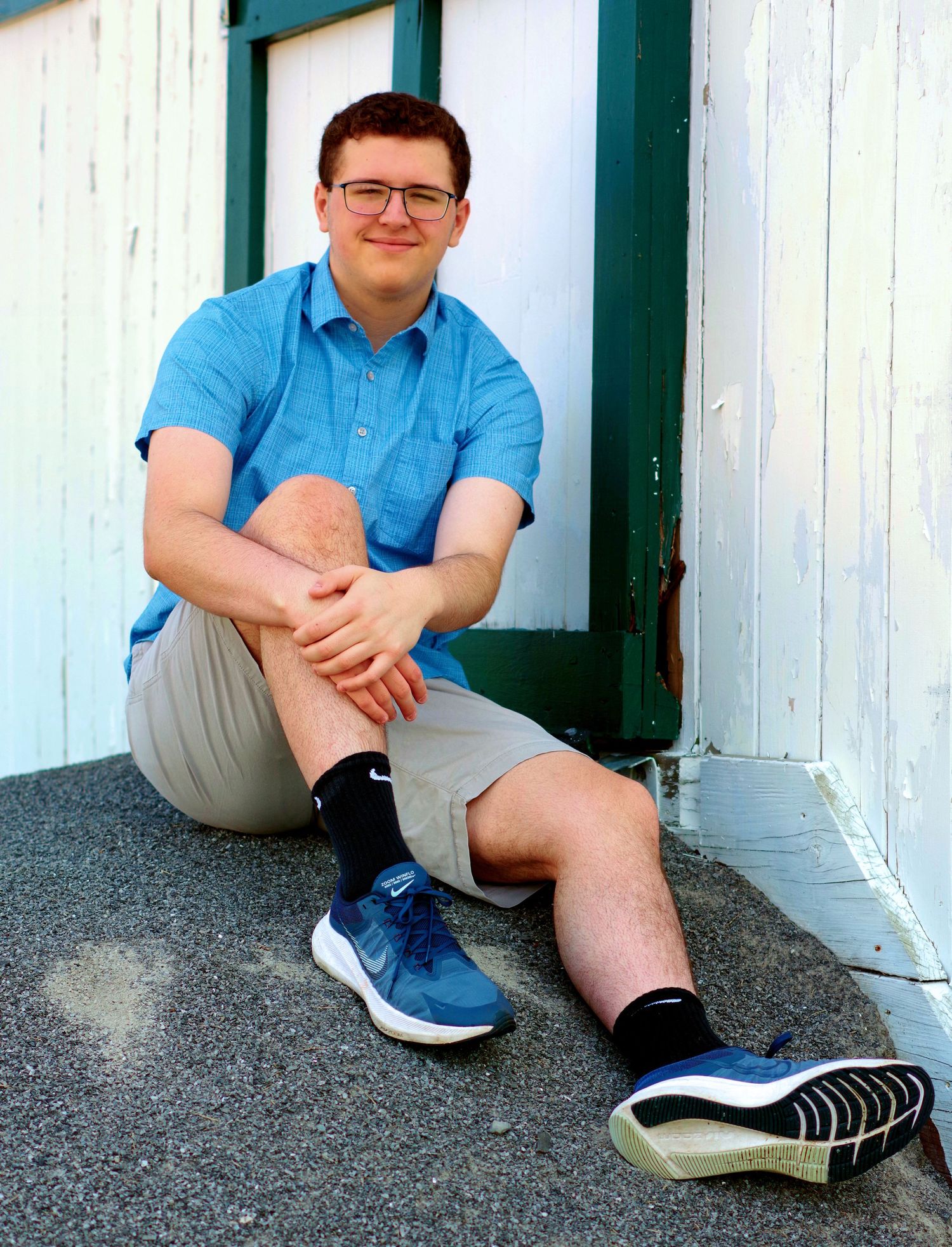 Casual seated portraits against white wall wearing blue shirt and khaki shorts.
