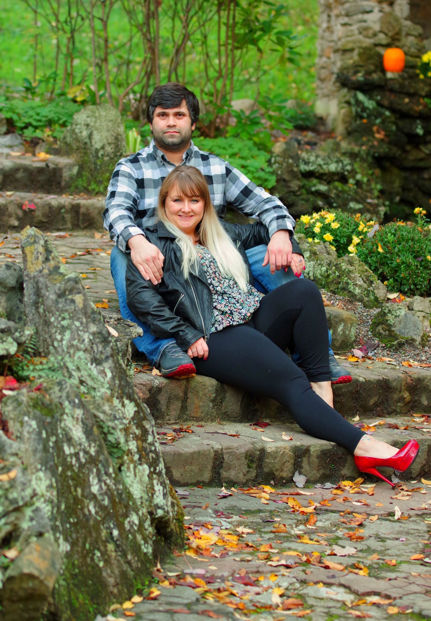 Couple sitting together on stone steps surrounded by autumn leaves and lush greenery in an outdoor garden setting.