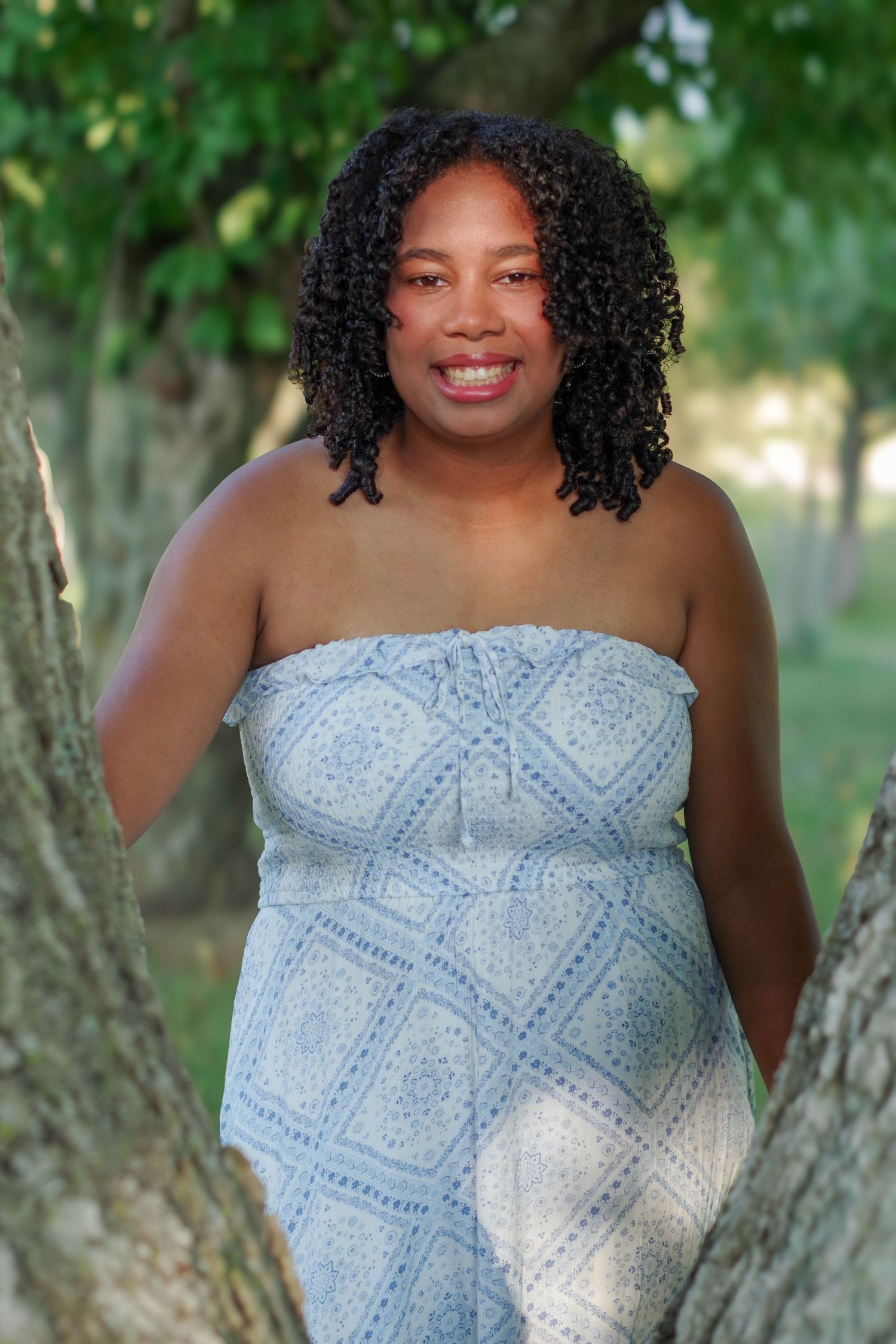 Person in blue patterned strapless dress poses between trees in sunny outdoor setting.
