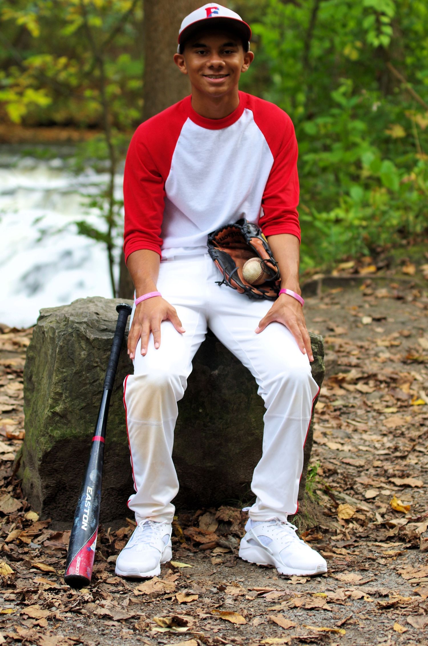 Baseball player in red and white uniform sitting with bat and glove by waterfall.