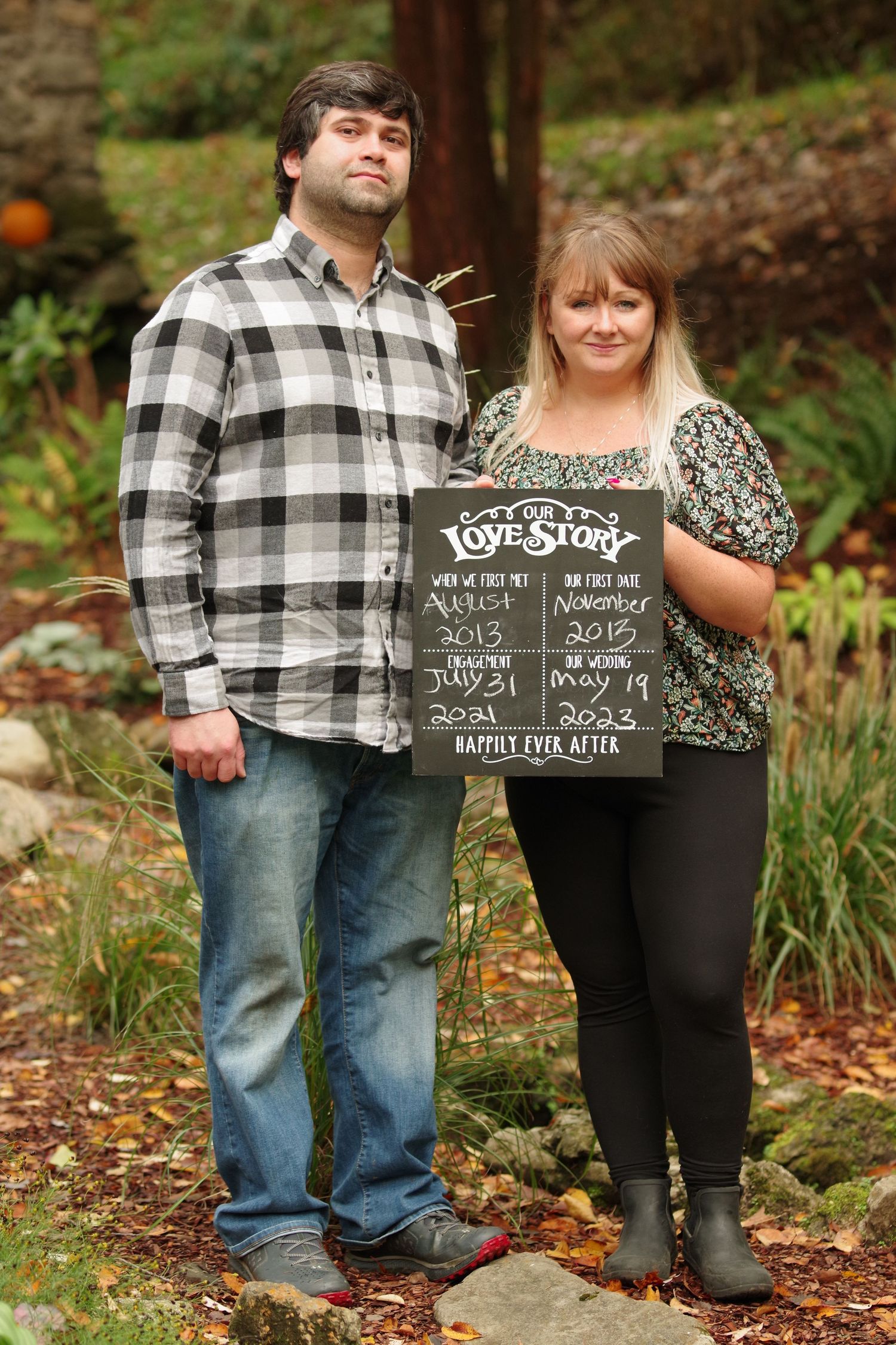 Couple holding a love story chalkboard sign with milestone dates in an autumn garden setting.