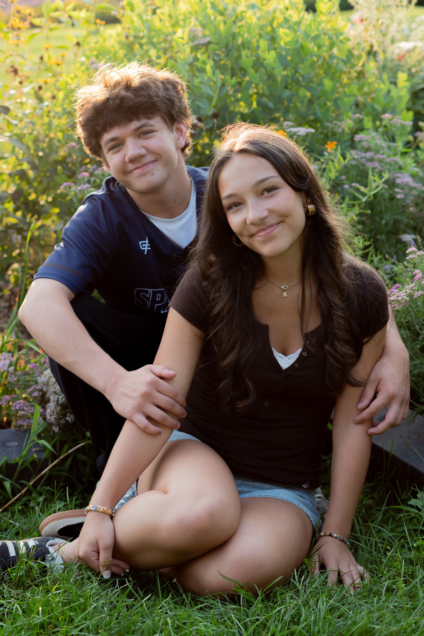 Two teenagers smiling and posing together outdoors in a garden setting on a sunny day.