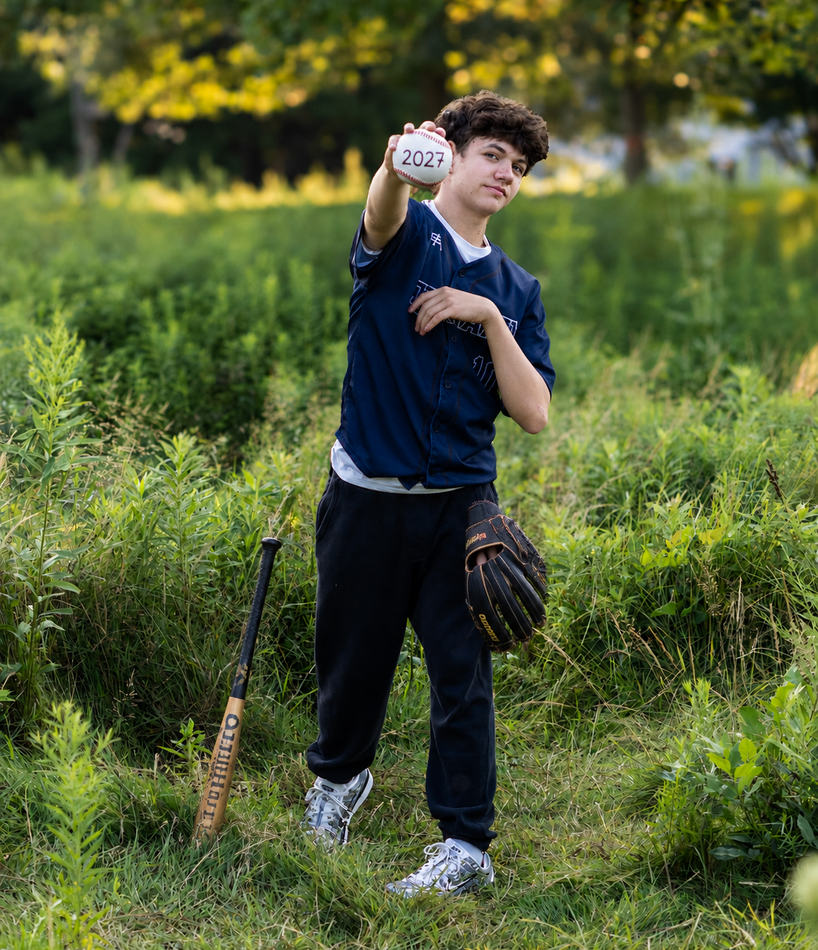 Teen baseball player in navy uniform holds a 2021 ball, with bat and glove, posing outdoors in a lush green field.