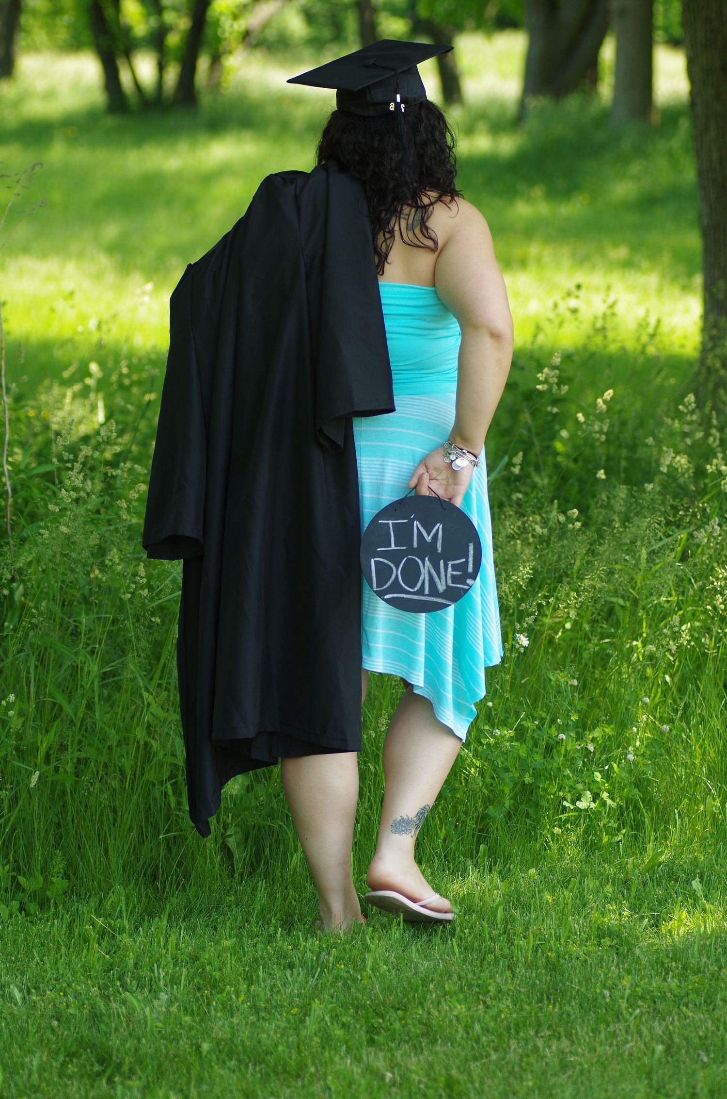 Graduate in turquoise dress and graduation attire holds chalkboard sign in outdoor garden setting.