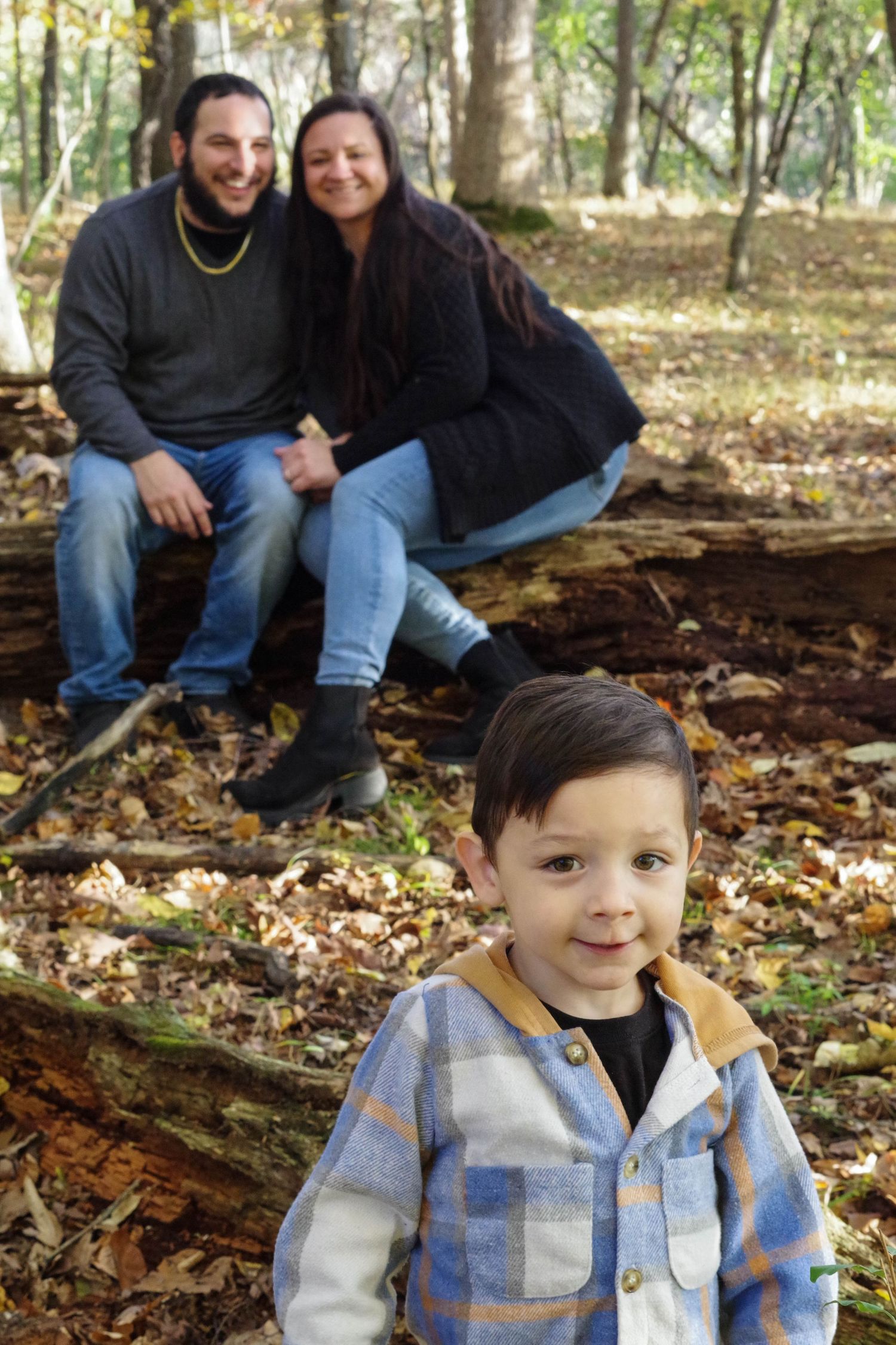 Family poses for outdoor portrait on fallen logs in wooded autumn setting.