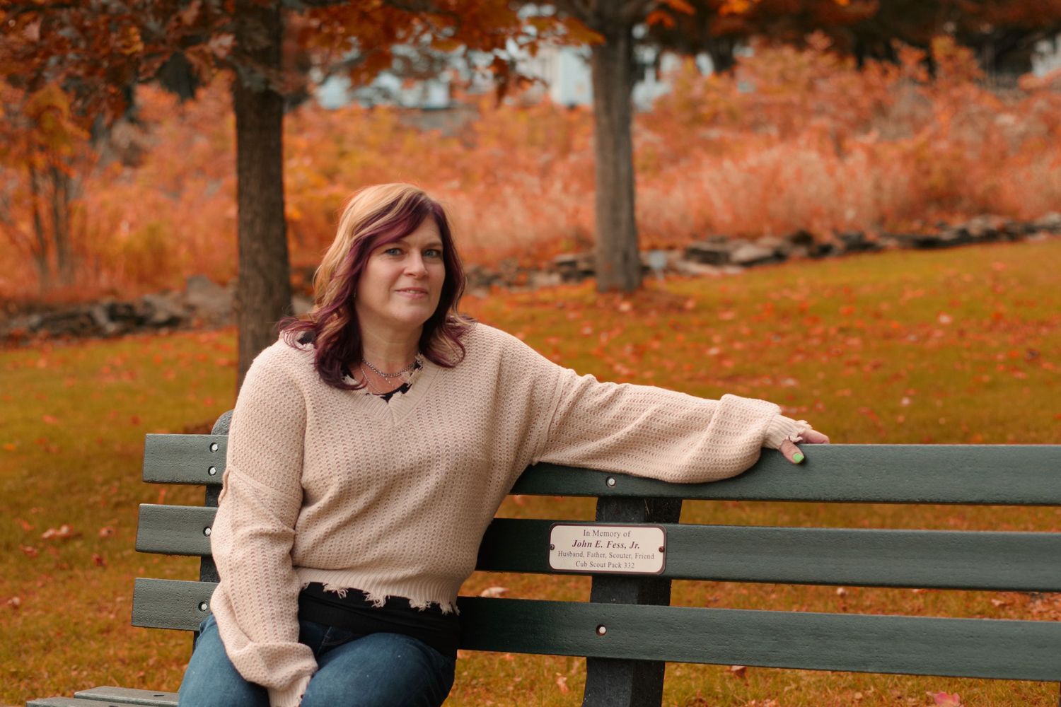 Person in beige sweater sits casually on park bench surrounded by fall foliage.