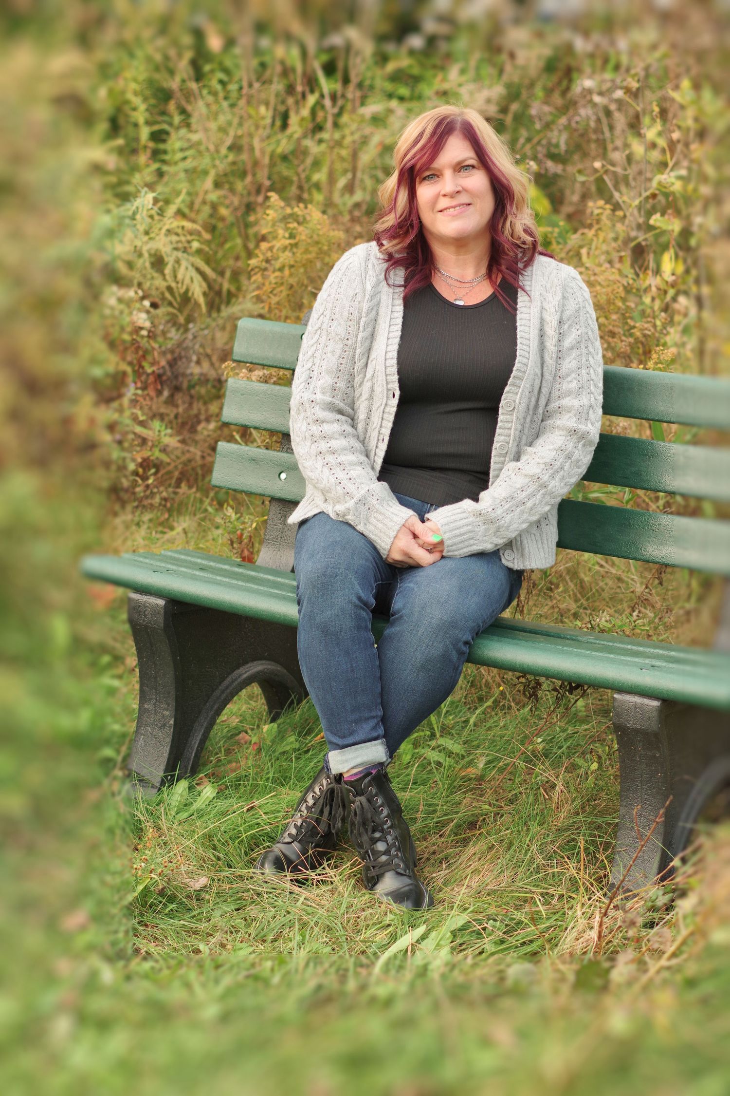 Person in cozy sweater and combat boots sits on green bench surrounded by tall grass.
