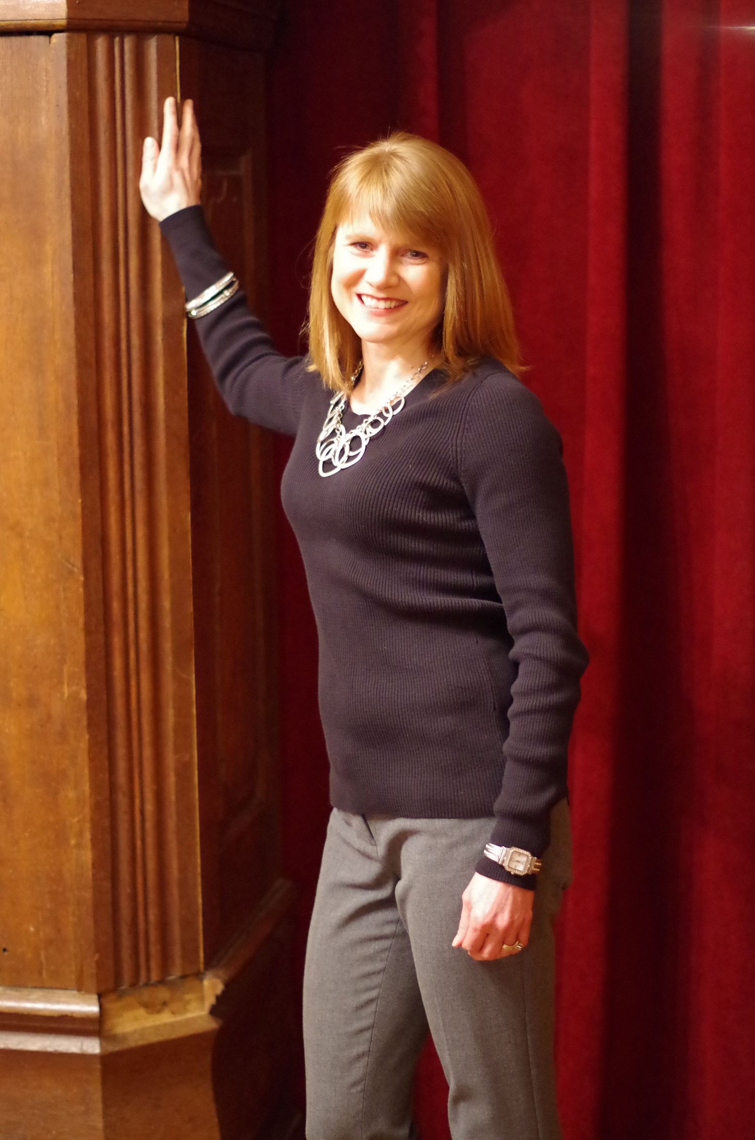 Full body shot with raised hand wearing dark sweater and grey pants against red curtain background.