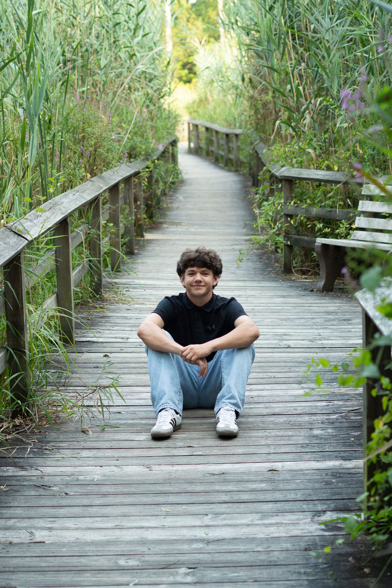 Teen boy sitting cross-legged on a wooden boardwalk path surrounded by tall green reeds, smiling at camera.