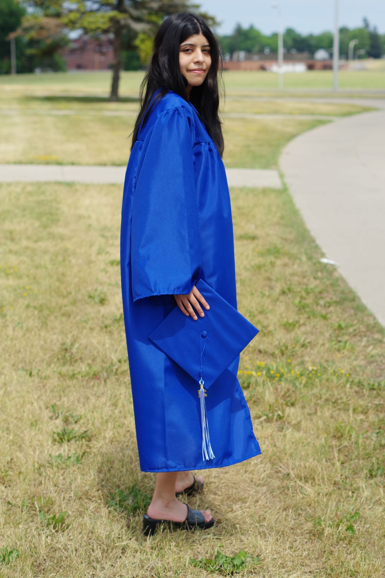 Graduate in flowing blue graduation gown stands on campus sidewalk on sunny day.