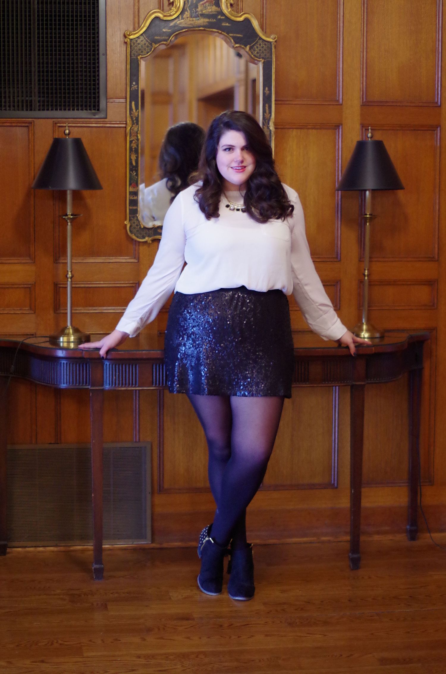 A person poses against a wooden wall with mirror and lamps wearing white top and sparkly skirt.