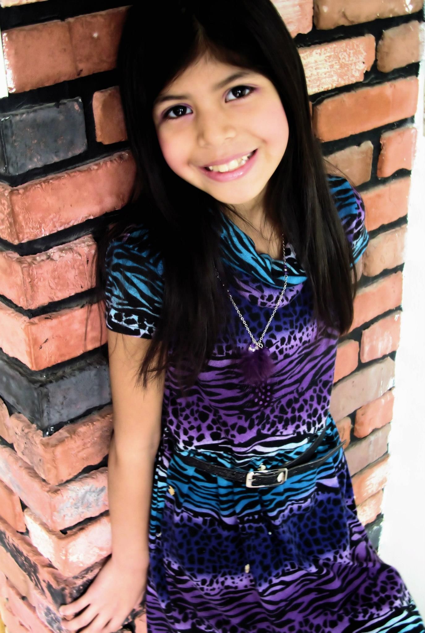 A person in a blue and purple patterned dress poses against a brick wall.