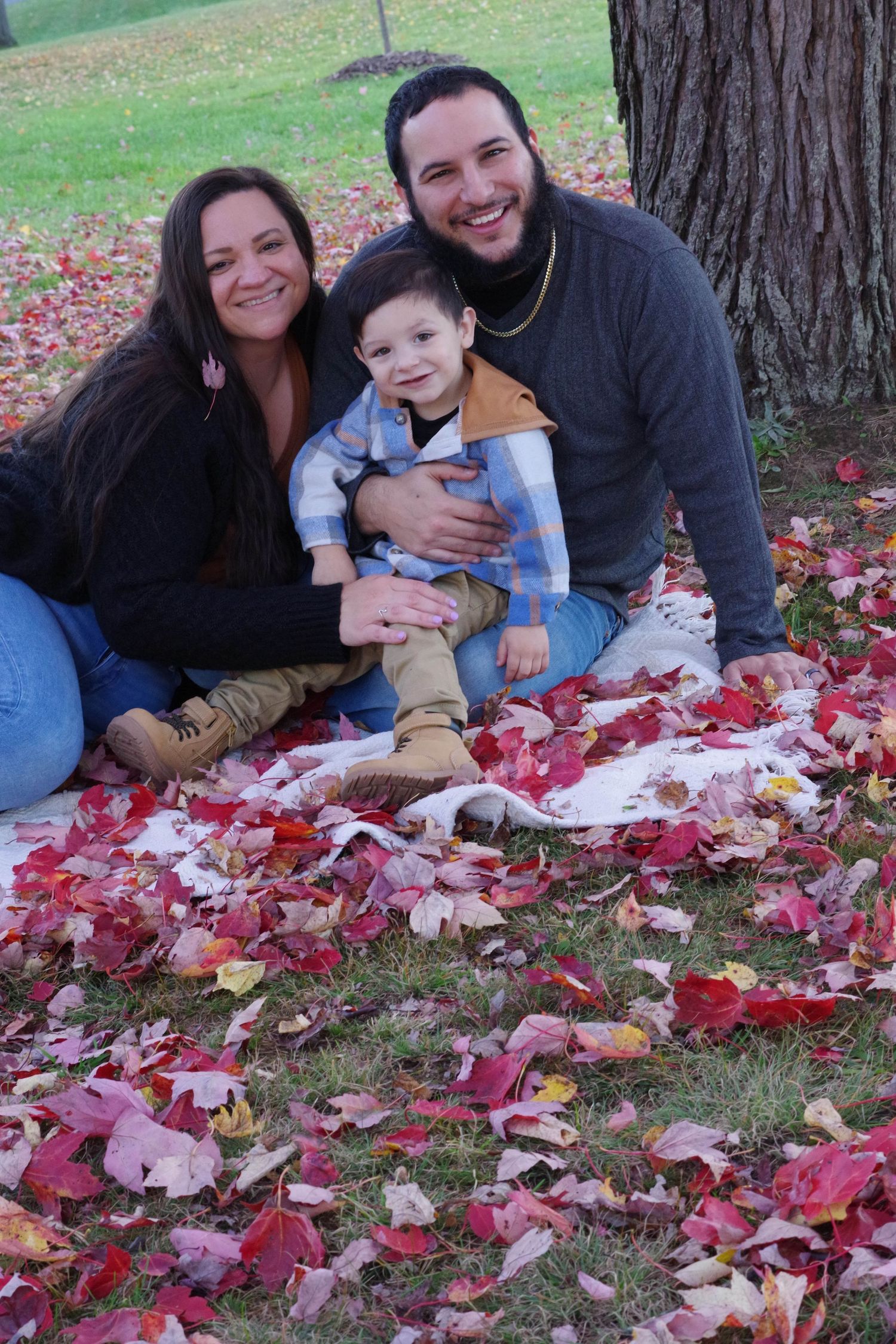 Family sits on blanket surrounded by vibrant red fall leaves in park setting.