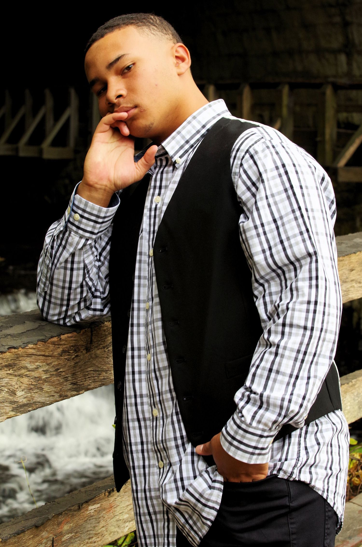 A person in checkered shirt and black vest poses thoughtfully with hand near face.